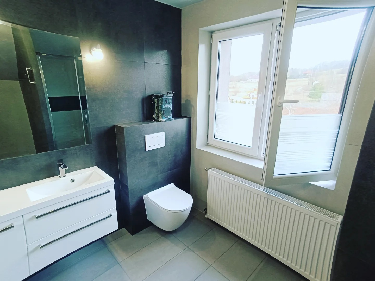 Nowoczesna łazienka z szarymi płytkami, białym umywalką z szafką, podwieszanym WC i otwartym oknem z widokiem na zewnątrz, dekorowana retro radiem na półce.