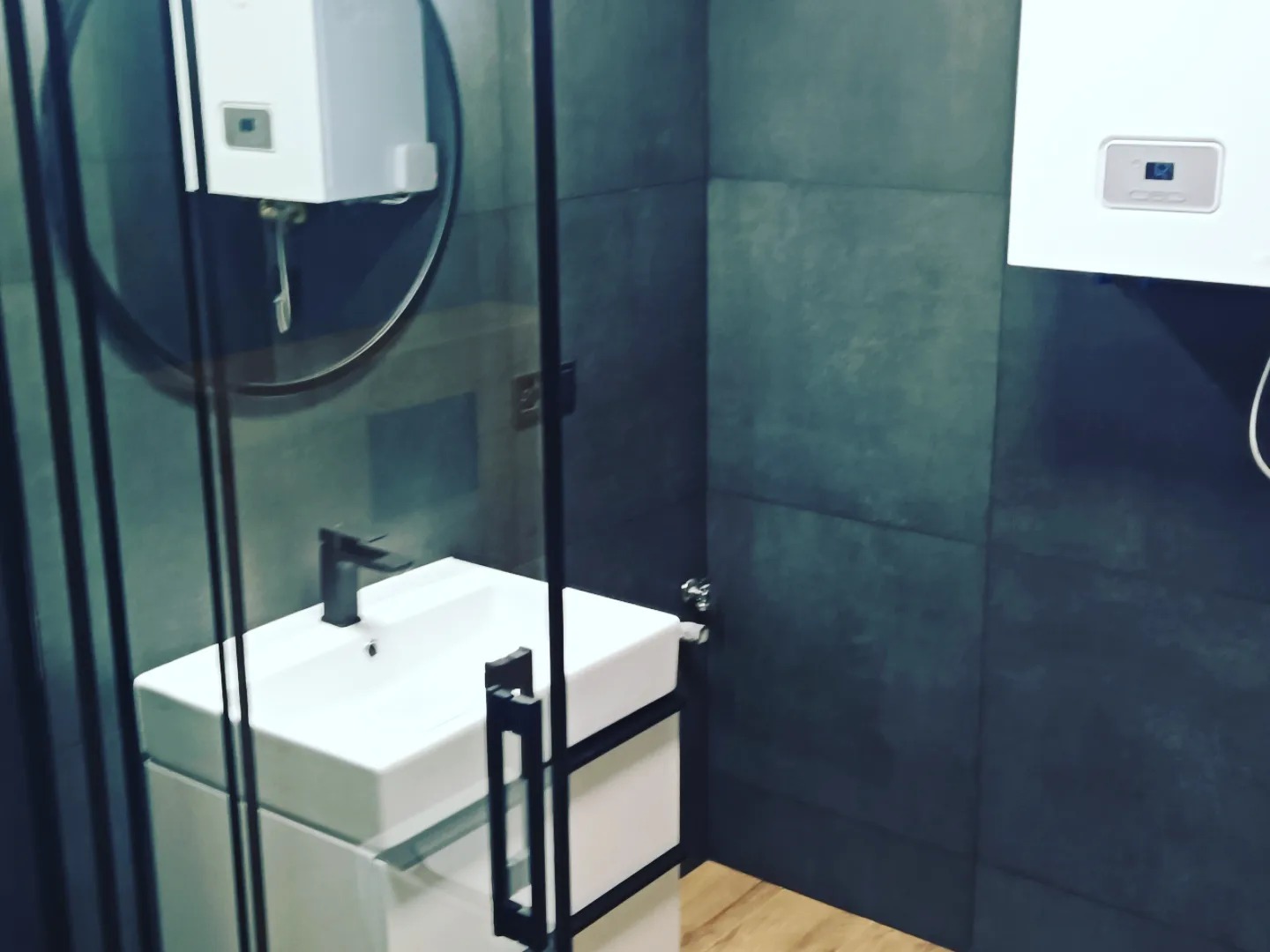 Nowoczesna łazienka z kwadratową umywalką nablatową, baterią w kolorze antracytu, okrągłym lustrem i szarymi płytkami wielkoformatowymi na ścianie, widoczny fragment kabiny prysznicowej ze...