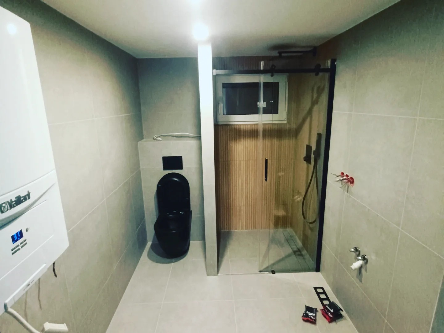 Wykończona łazienka z kabiną prysznicową typu walk-in z czarną ramą, wiszącą czarną toaletą i grzejnikiem Vaillant na ścianie z szarymi płytkami. Na podłodze leżą narzędzia.