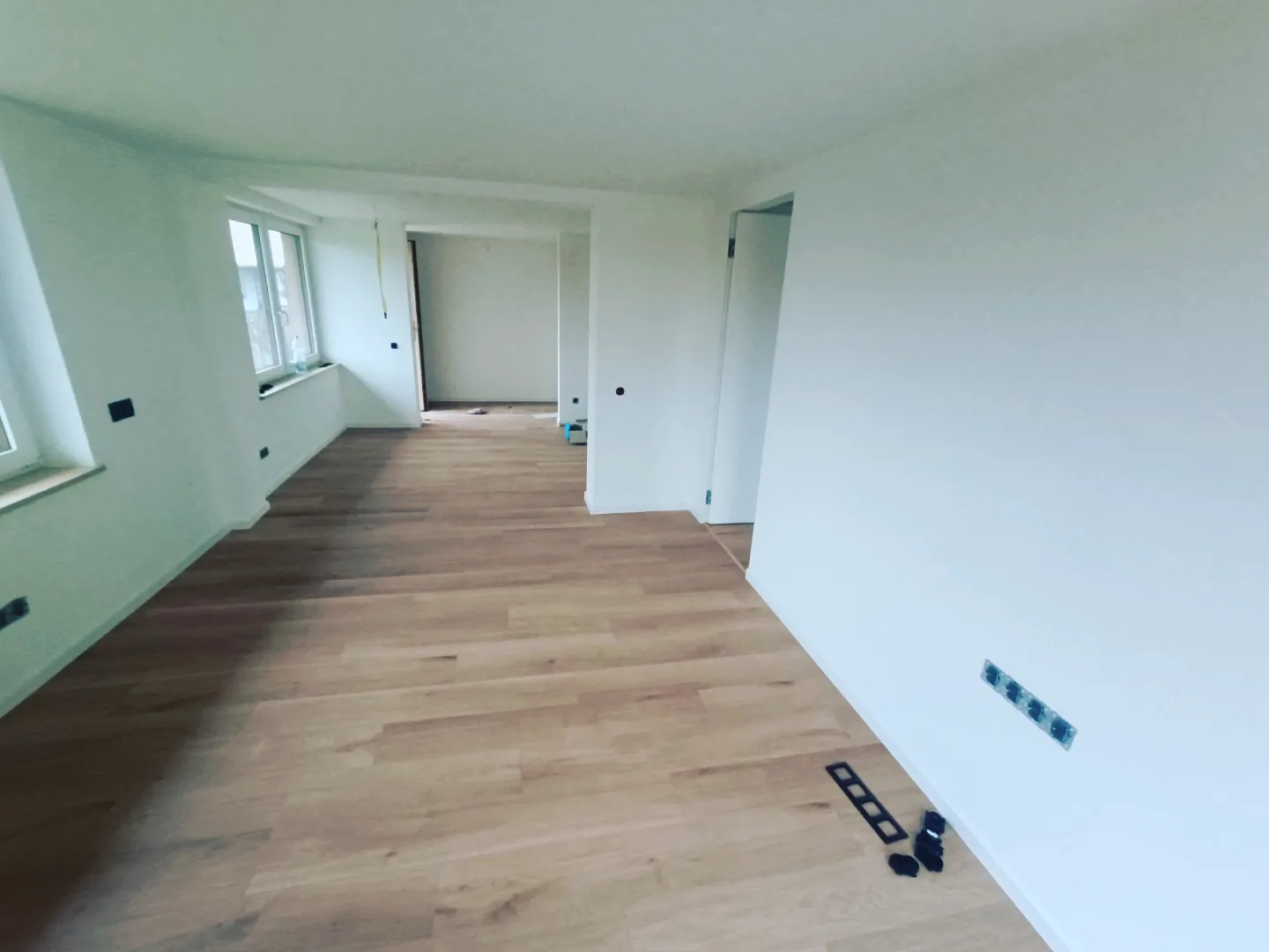 Jasne, nowocześnie wykończone wnętrze z drewnianą podłogą, świeżo pomalowanymi białymi ścianami z widocznymi gniazdkami elektrycznymi i elementami montażowymi leżącymi na podłodze.