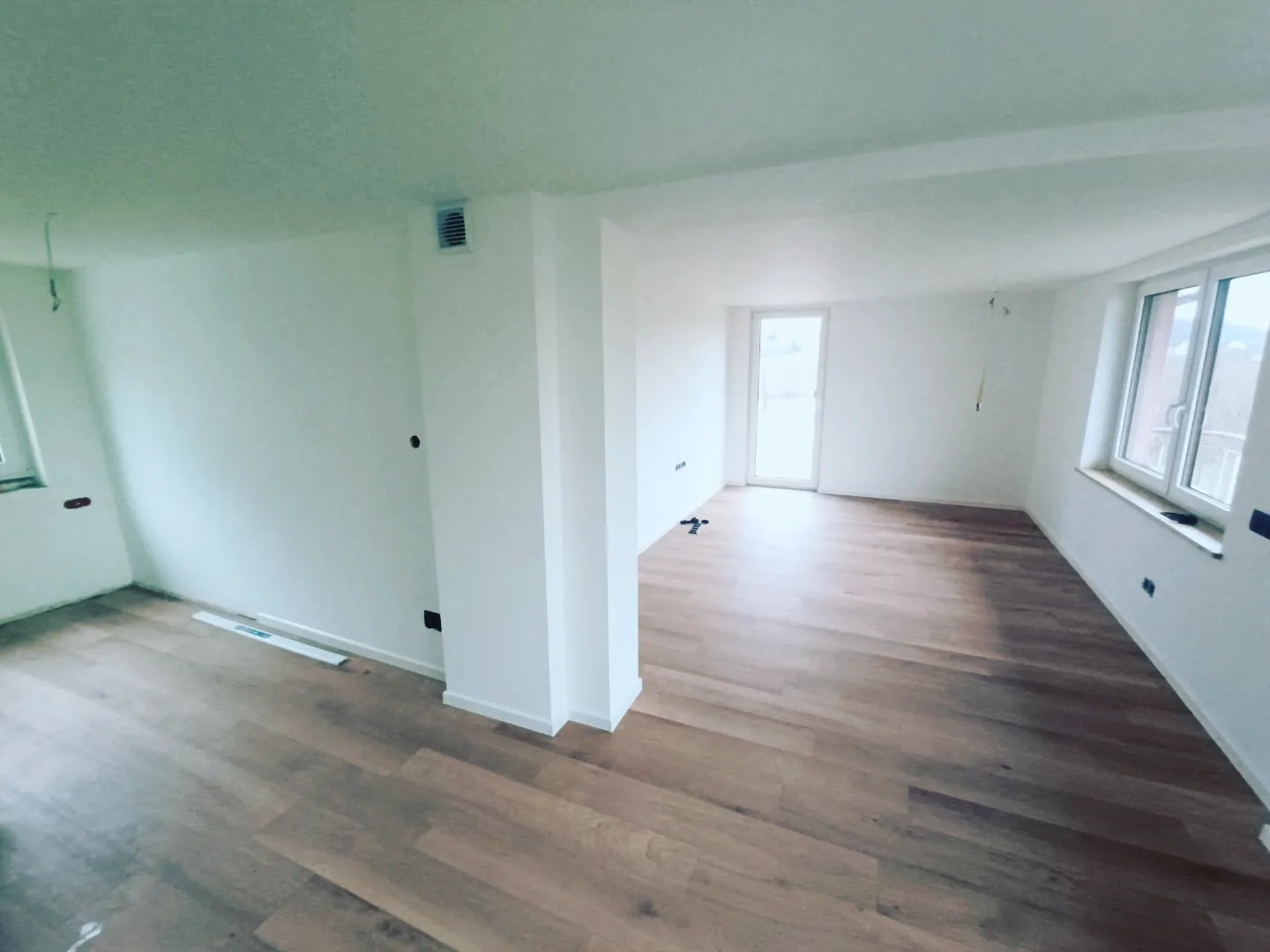 Jasne, przestronne wnętrze z nowo ułożoną podłogą z paneli w odcieniach dębu, świeżo pomalowane na biało ściany, widoczne punkty elektryczne i kratka wentylacyjna.