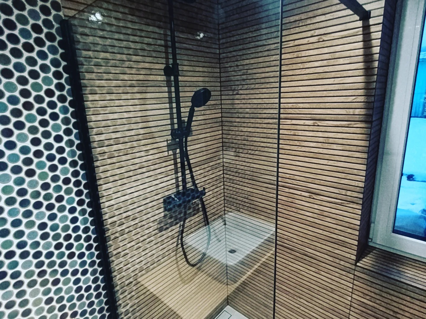 Nowoczesna kabina prysznicowa z deszczownicą w czarnym kolorze, wyłożona płytkami imitującymi drewniane lamele i mozaiką z okrągłych płytek w odcieniach niebieskiego i zielonego.