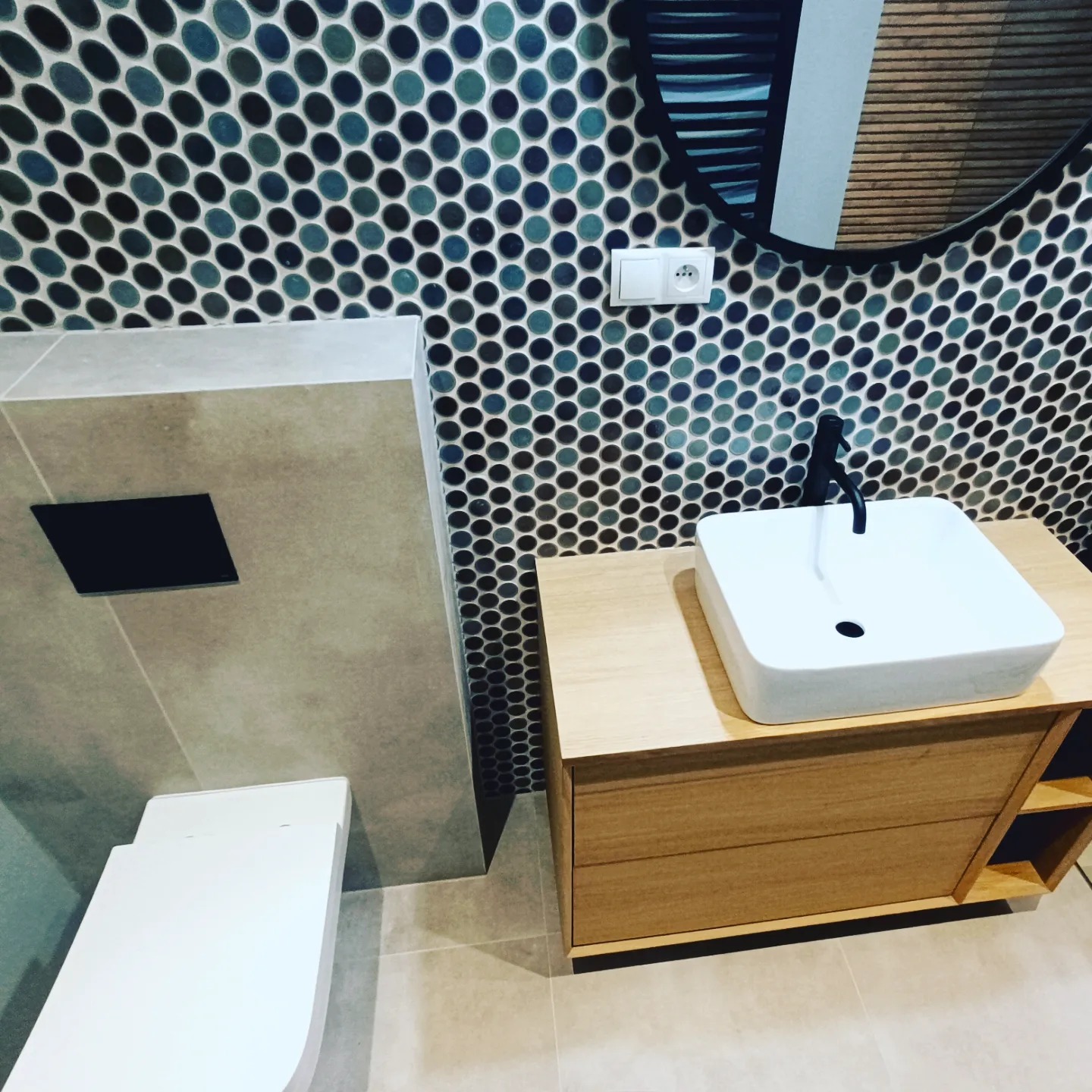 Nowoczesna łazienka w Bochni z mozaikową ścianą w odcieniach niebieskiego i czarnego, minimalistyczną białą umywalką nablatową z czarną armaturą i drewnianą szafką pod umywalką, oraz podwieszaną...