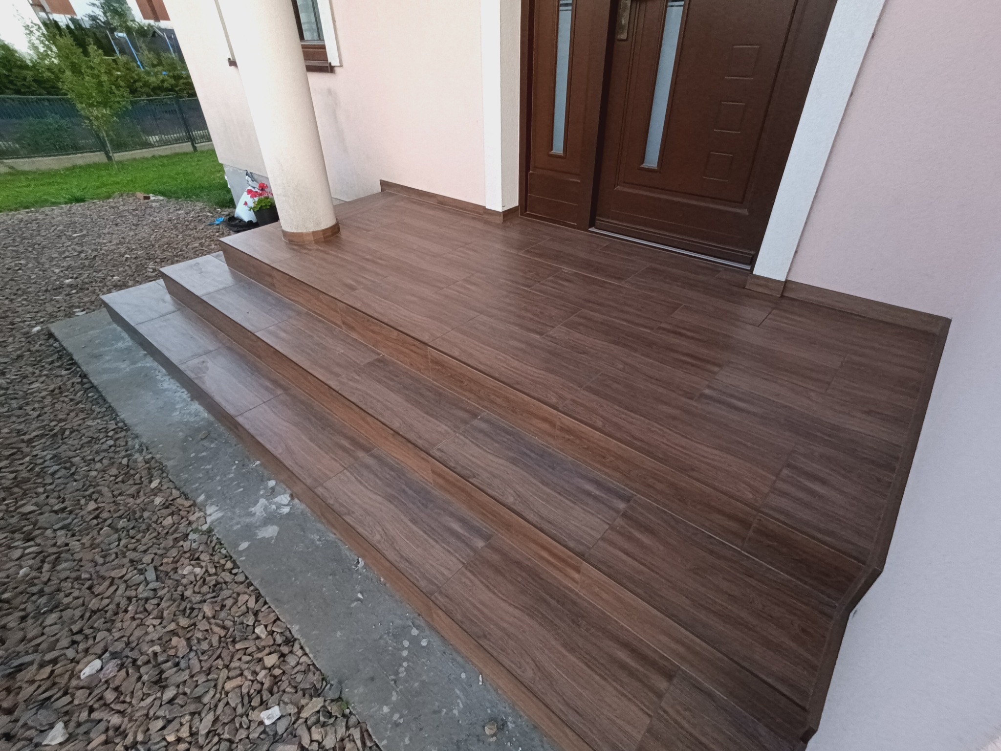Wykończone schody wejściowe do domu z brązowych płytek imitujących drewno, prowadzące do ciemnobrązowych drzwi wejściowych, z widoczną kolumną i fragmentem elewacji w tle.