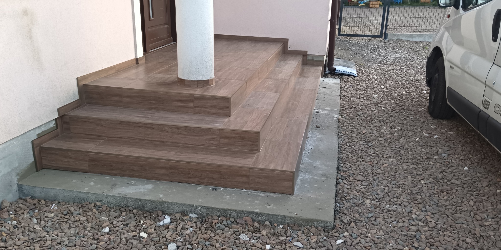 Wykończone schody wejściowe do domu z brązowych płytek imitujących drewno, otoczone szarym żwirem, z widoczną białą kolumną i fragmentem zaparkowanego białego samochodu.
