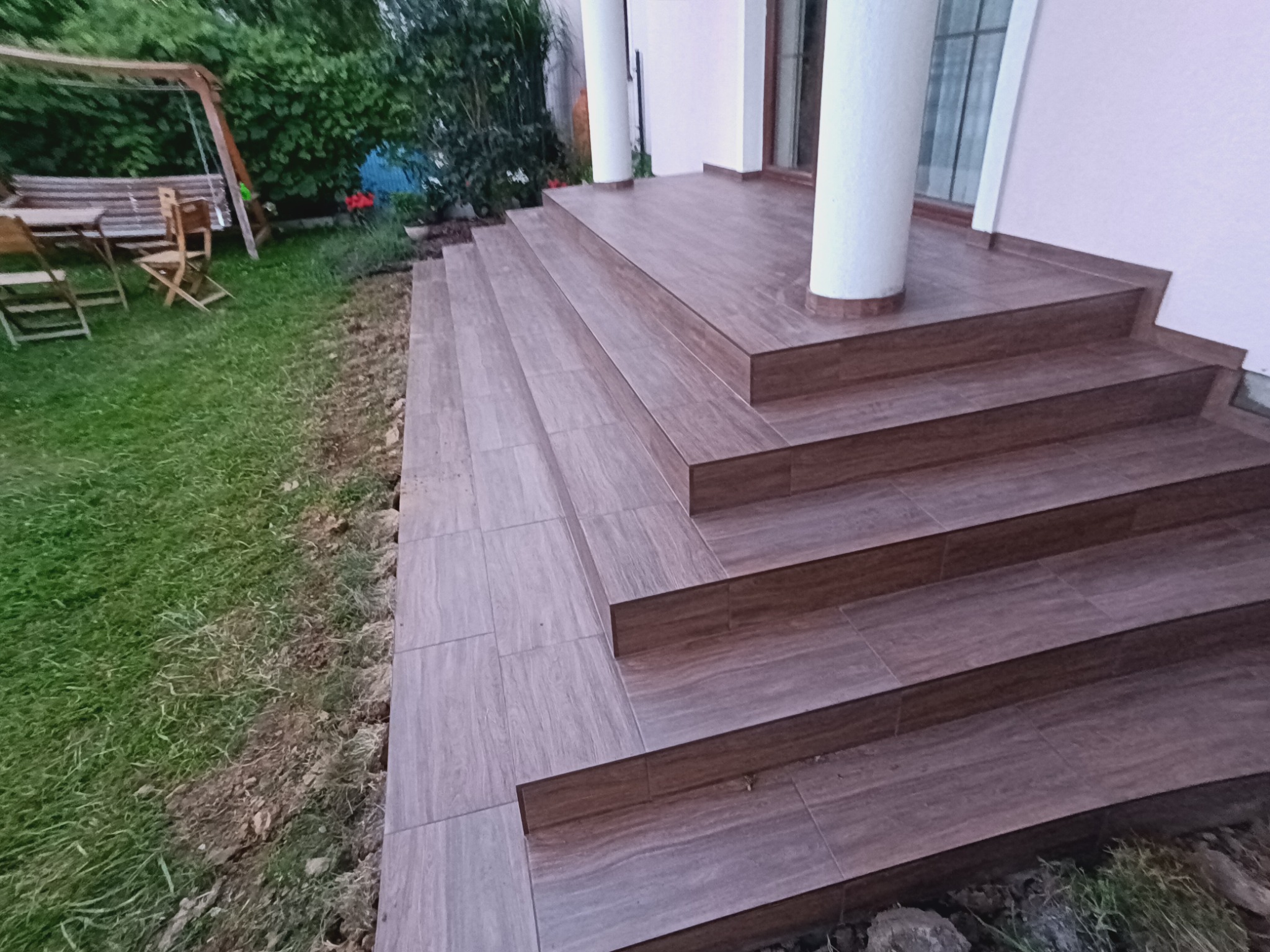 Zewnętrzne schody i taras wyłożone brązowymi płytkami imitującymi drewno, z widocznymi słupami i fragmentem trawnika po bokach.