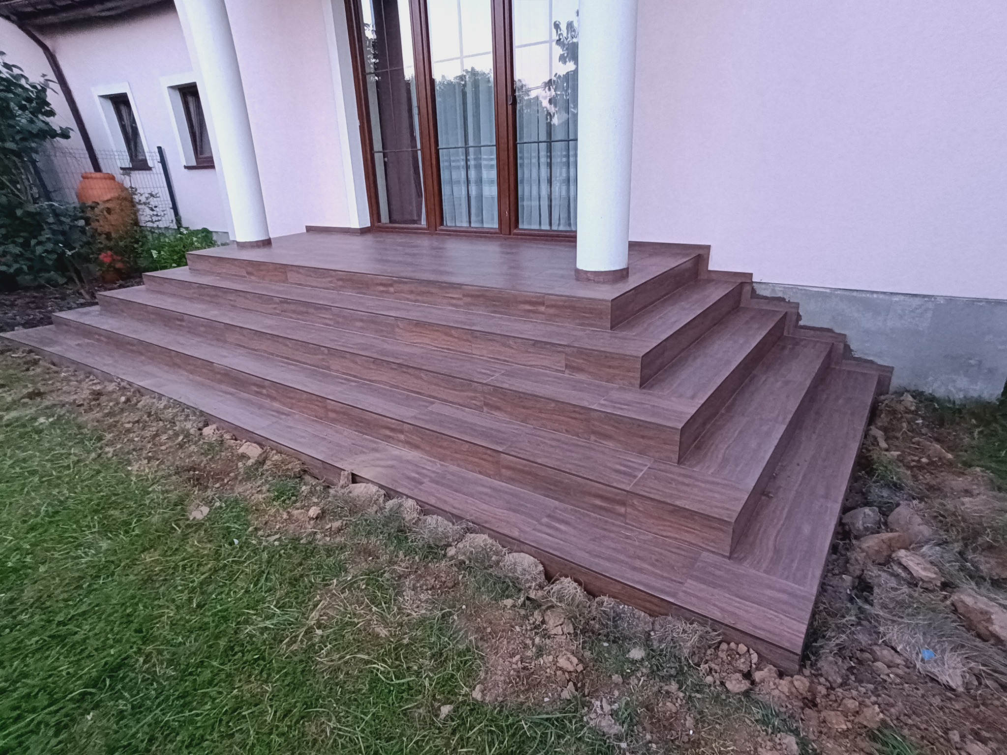 Zewnętrzne schody wejściowe do domu wyłożone brązowymi płytkami imitującymi drewno, z widocznymi fugami, prowadzące do drzwi z kolumnami, otoczone trawnikiem i ziemią.