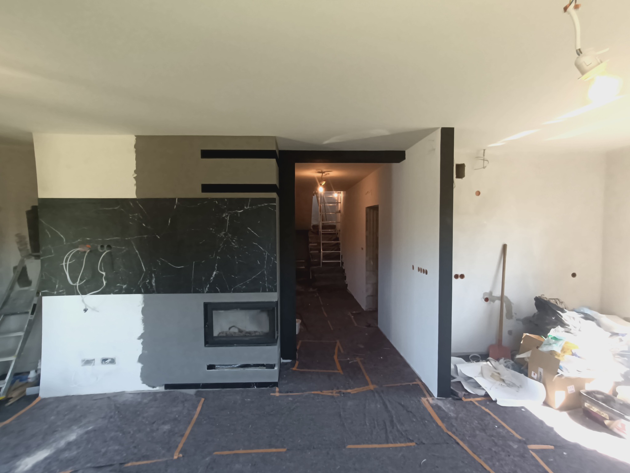 Wnętrze w trakcie remontu: częściowo wykończony kominek z czarnym marmurem, fragment ściany z nałożoną gładzią, zabezpieczona podłoga, widoczne narzędzia i drabina.