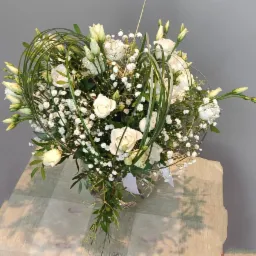 Biały bukiet z róż, frezji i gipsówki w szklanym wazonie, z dekoracyjnymi zielonymi pędami ułożonymi w okrąg wokół kwiatów, na tle szarej ściany i blatu z wzorem cieni.