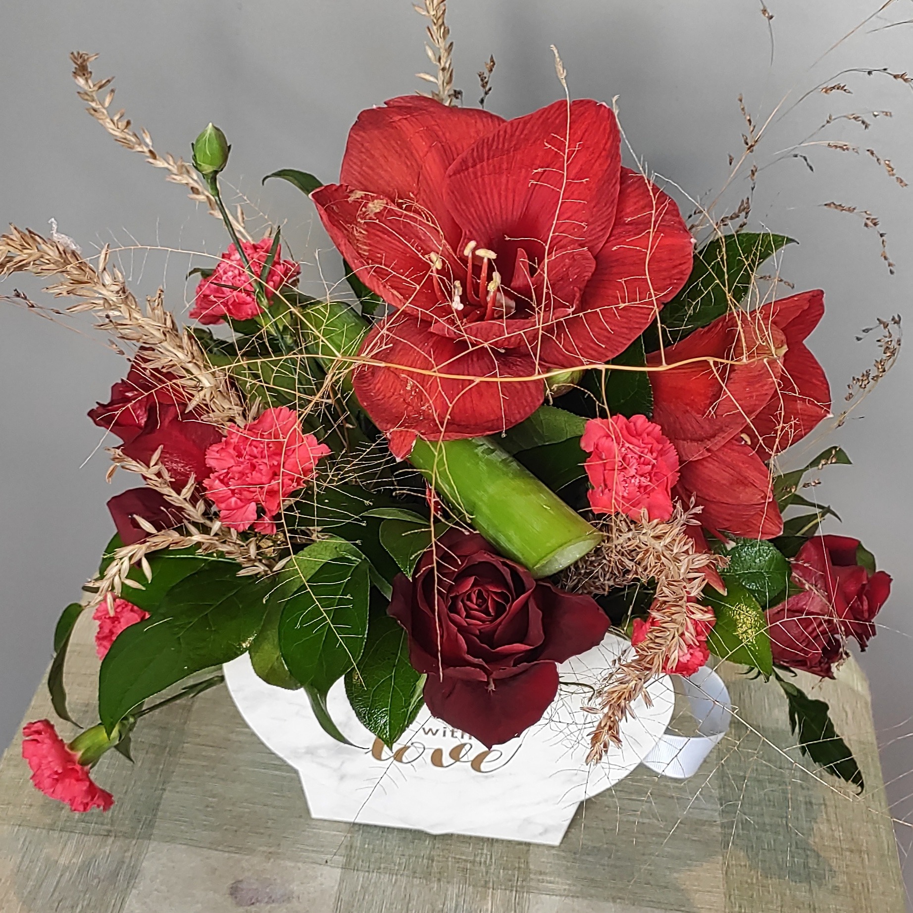 Kompozycja z czerwonych amarylisów, goździków i róży w białym wazonie z napisem 'with love', uzupełniona suszonymi trawami i zielonymi liśćmi.