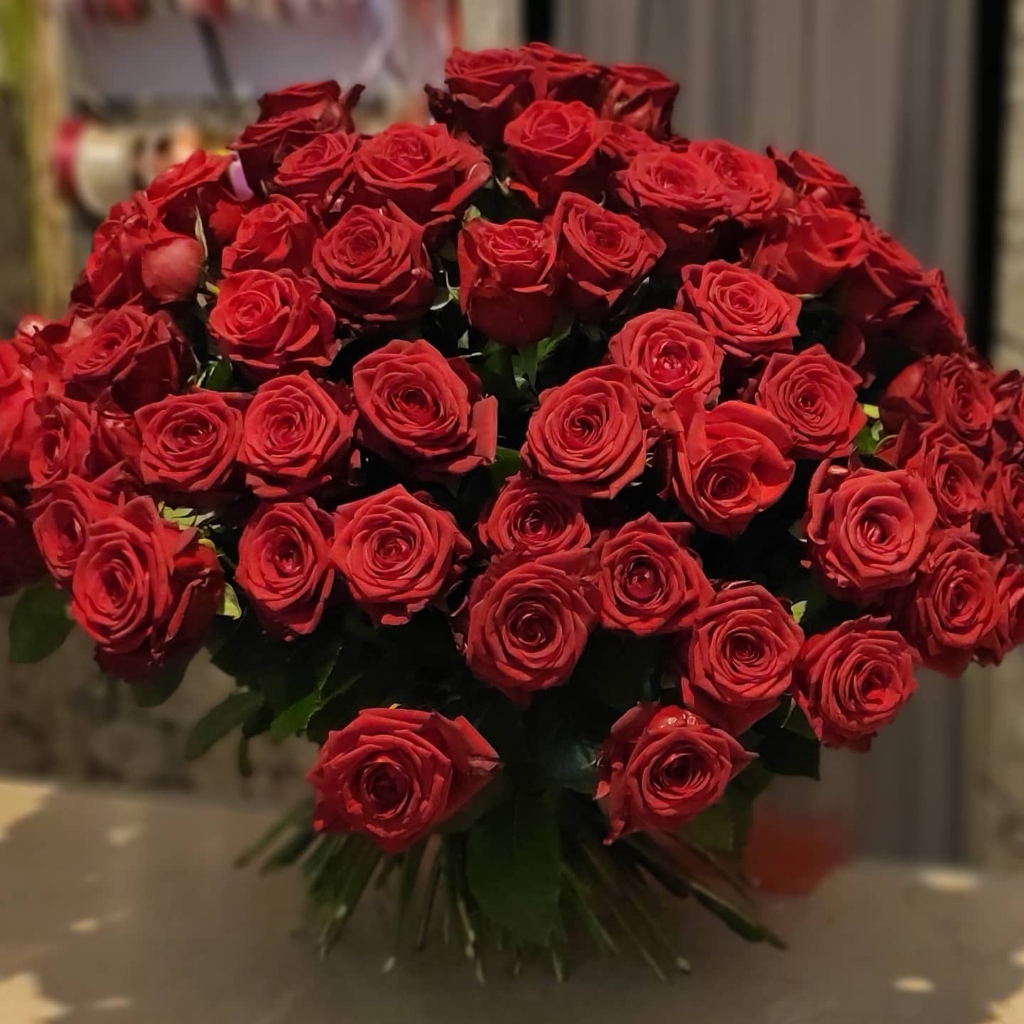 Duży bukiet intensywnie czerwonych róż, ciasno ułożonych, z widocznymi zielonymi łodygami u podstawy, na rozmytym tle wnętrza.