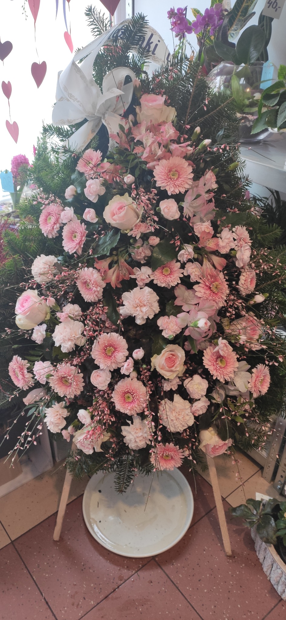 Kompozycja kwiatowa w odcieniach różu z gerberami, różami i drobnymi kwiatkami, umieszczona na drewnianym stojaku w kwiaciarni.