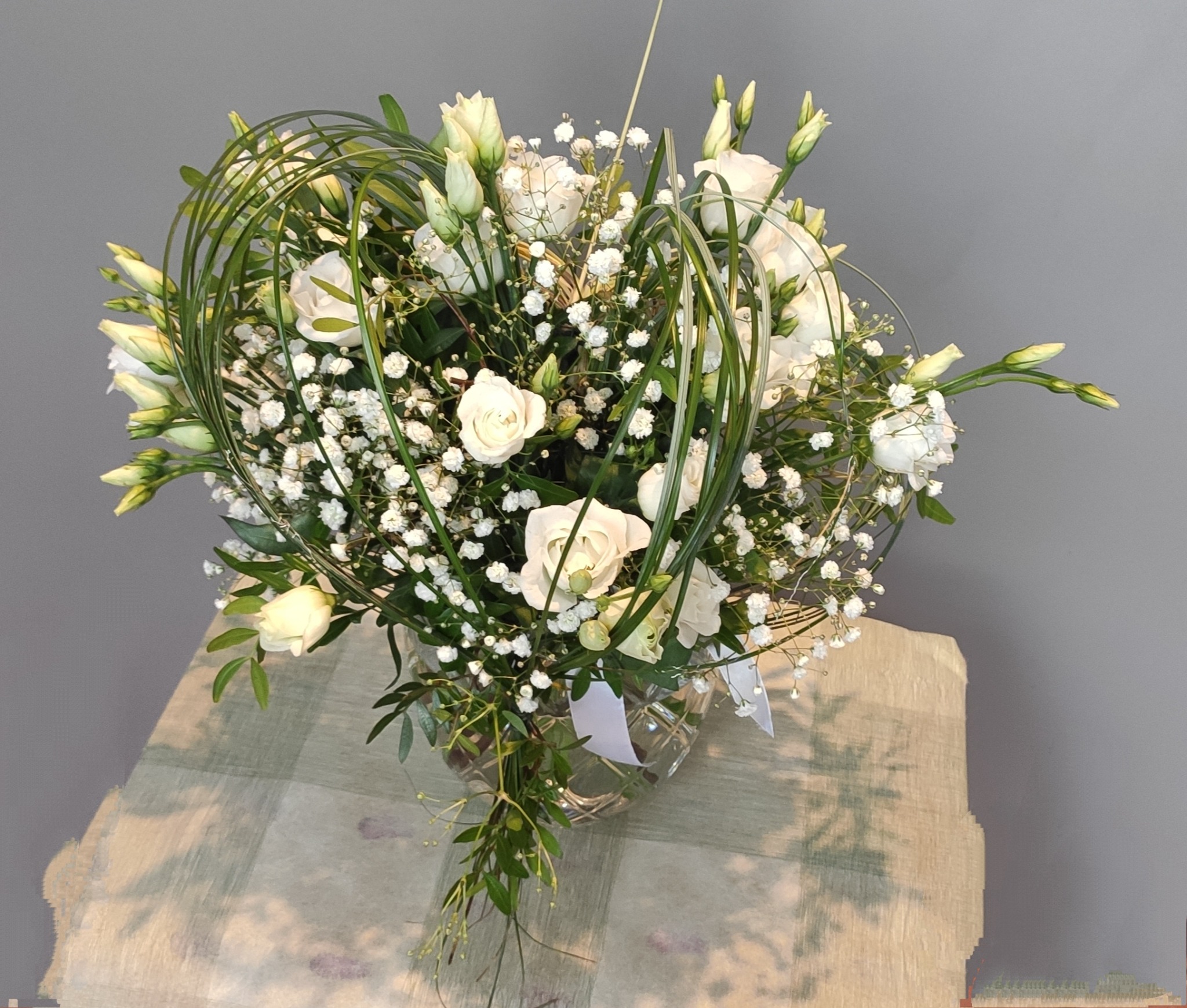 Biały bukiet z róż, frezji i gipsówki w szklanym wazonie, z dekoracyjnymi zielonymi pędami ułożonymi w okrąg wokół kwiatów, na tle szarej ściany i blatu z wzorem cieni.
