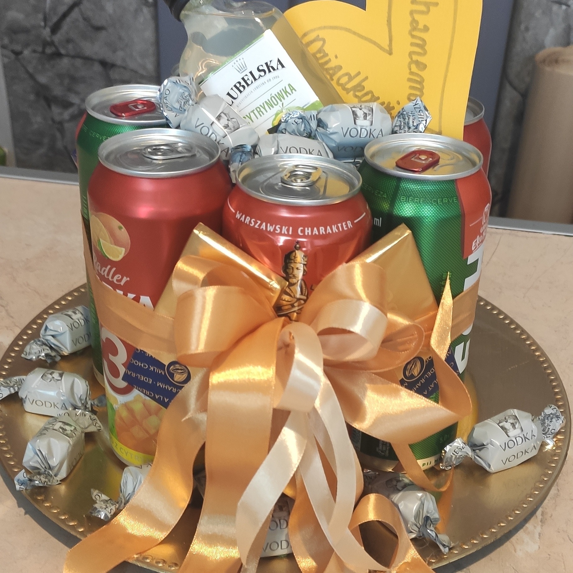 Dekoracyjny kosz prezentowy z piwami, wódką Lubelską, pralinkami Vodka i złotą wstążką na złotej tacy.