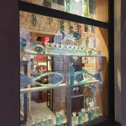 Ekspozycja sklepu z ręcznie robionymi szklanymi ozdobami: figury ryb, wazony i świeczniki na tle ceglanej ściany i widocznego wnętrza sklepu.