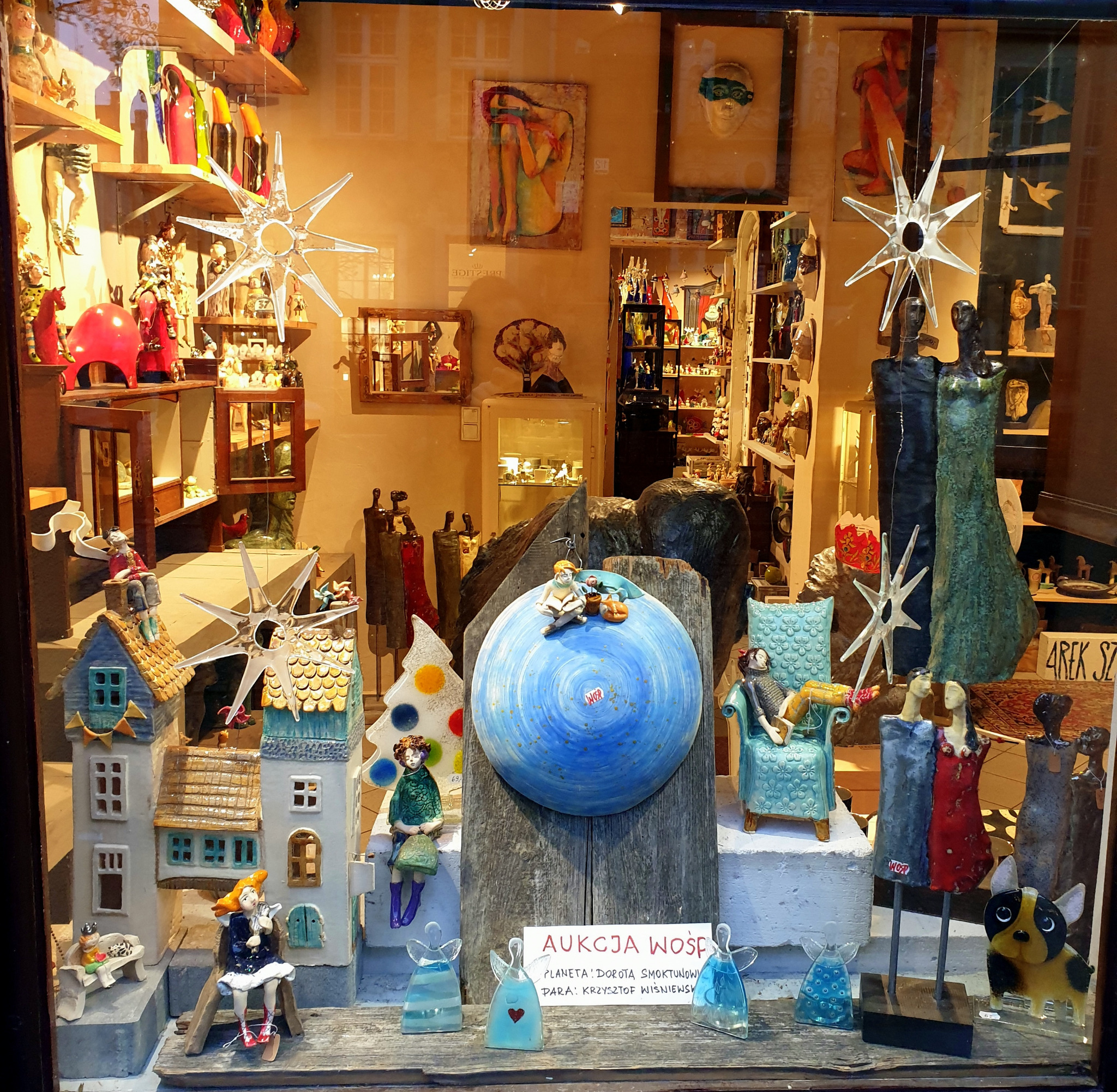 Ekspozycja w witrynie galerii sztuki: ceramiczne domki, szklane aniołki, figury ludzi i zwierząt, obrazy na ścianie, oświetlone gwiazdy, aukcja Woś na kartce.