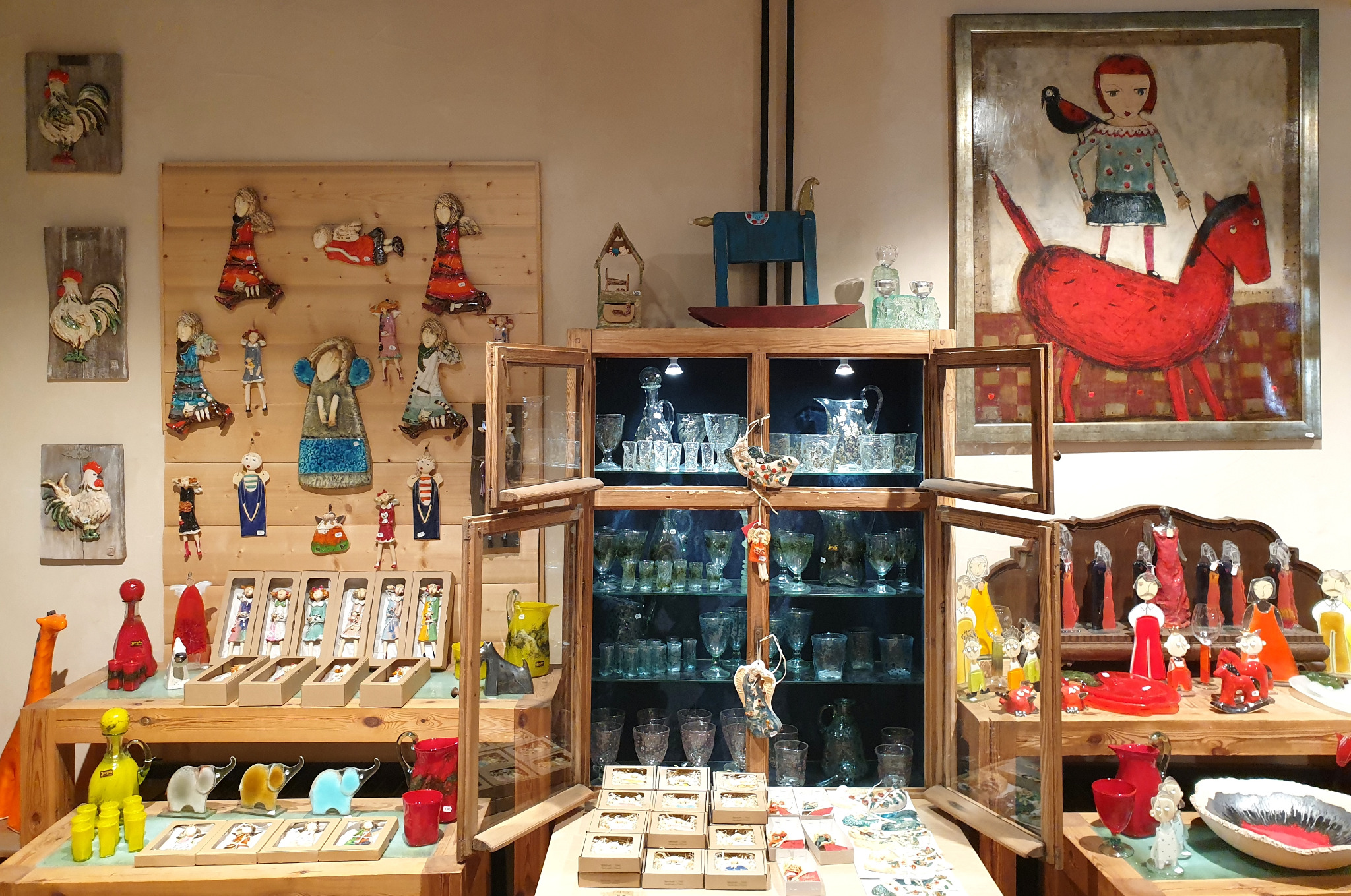 Wystawa rękodzieła artystycznego w sklepie: ceramiczne figurki, malowane obrazy, szklane naczynia i ozdoby.