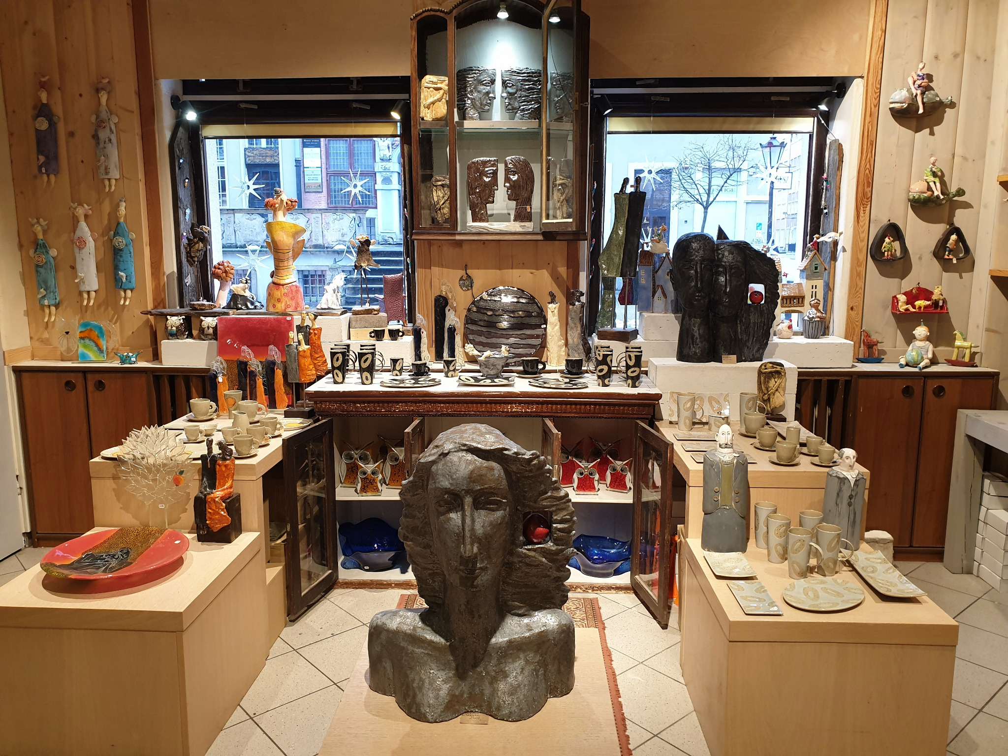 Wnętrze sklepu z rękodziełem artystycznym: ceramiczne rzeźby, naczynia i figurki na drewnianych półkach i w przeszklonej witrynie, widok z perspektywy klienta.