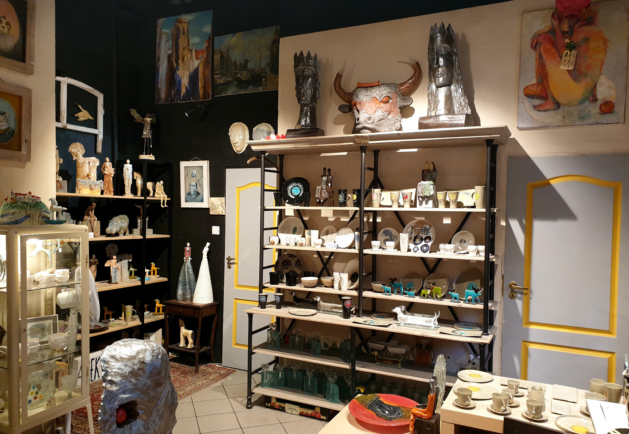 Wnętrze galerii sztuki z licznymi rzeźbami, obrazami i ceramiką na półkach i ścianach, w tym rzeźba głowy byka z rogami na górnej półce.
