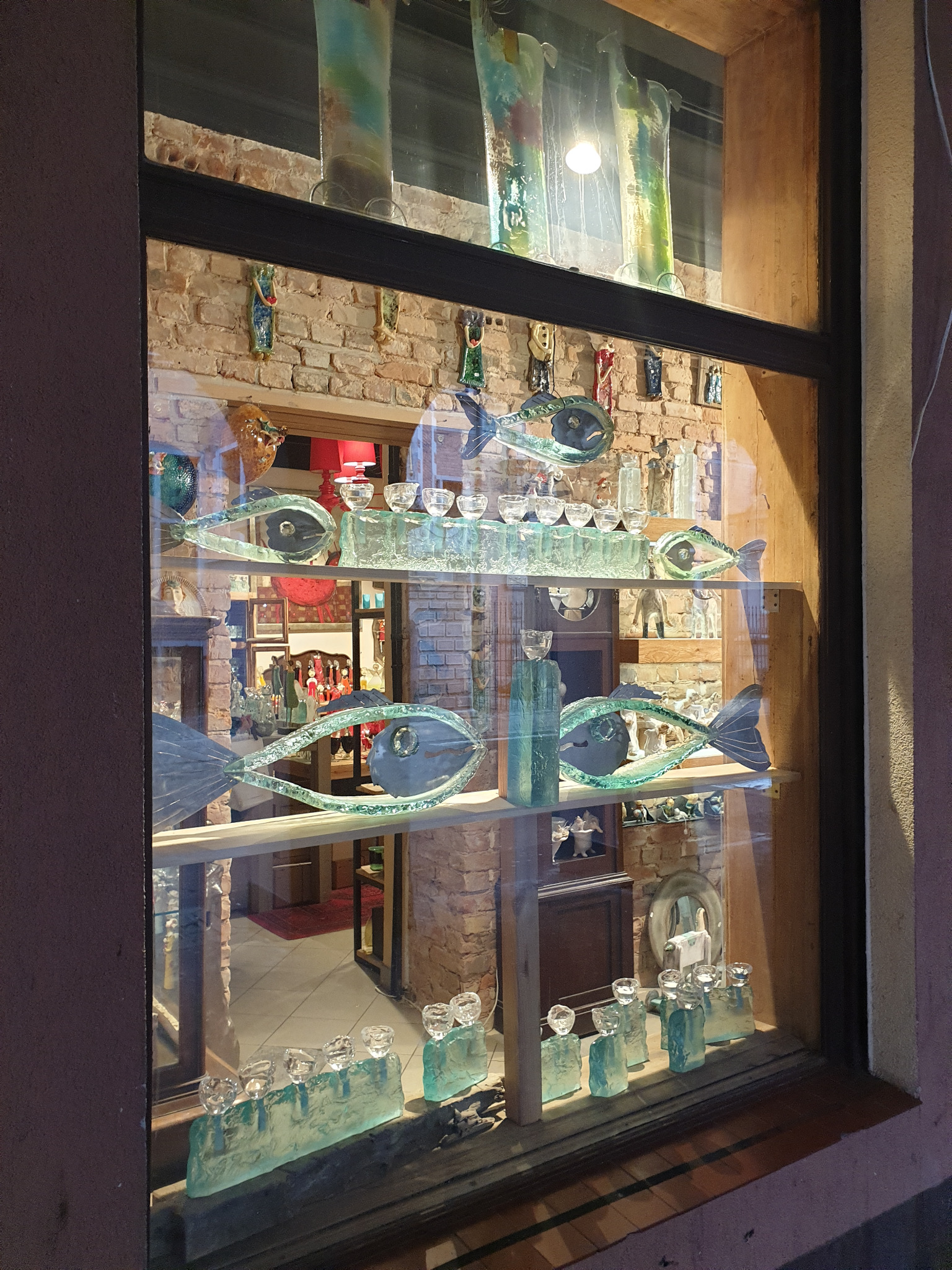Ekspozycja sklepu z ręcznie robionymi szklanymi ozdobami: figury ryb, wazony i świeczniki na tle ceglanej ściany i widocznego wnętrza sklepu.