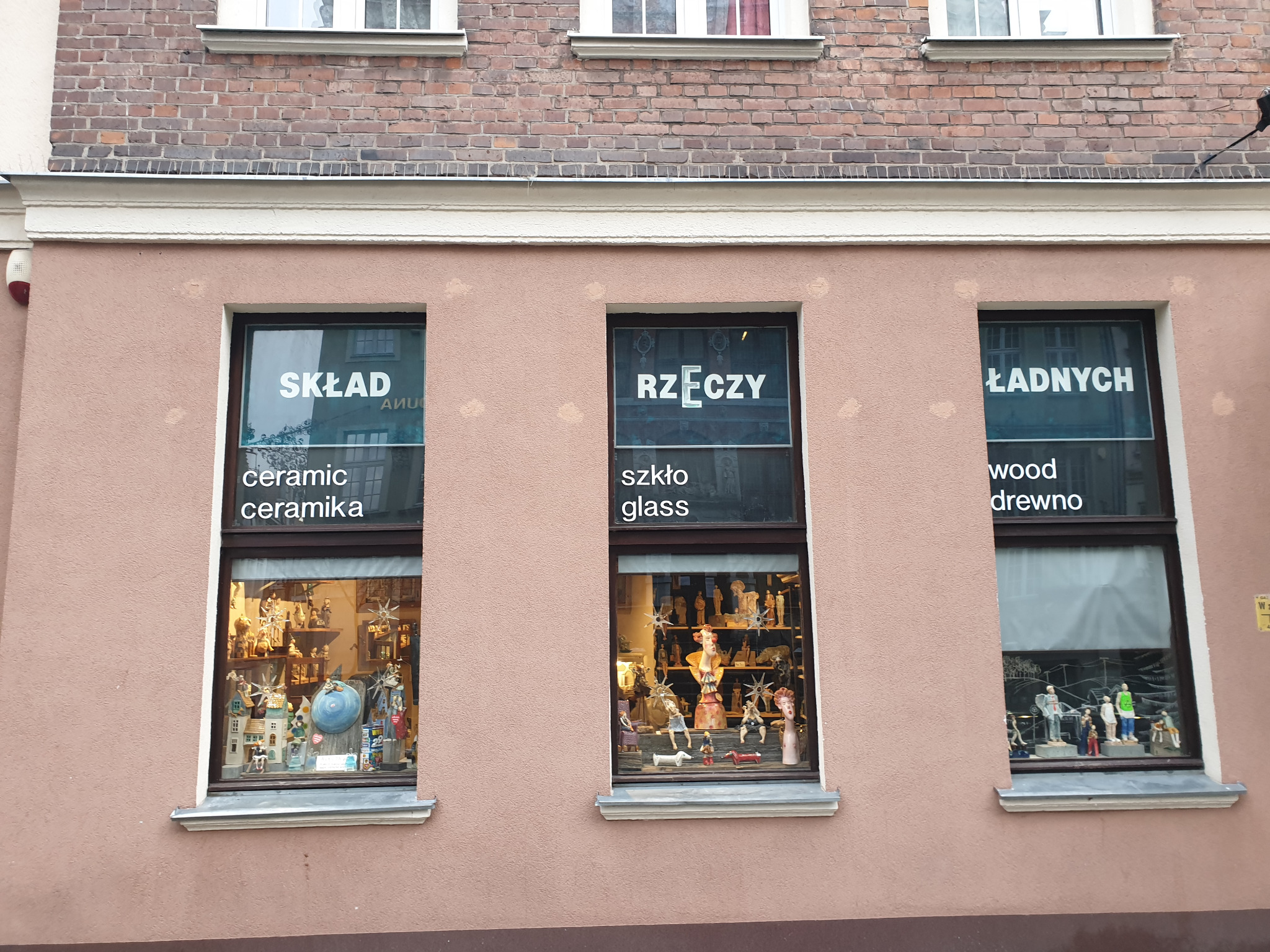 Fasada budynku z trzema oknami wystawowymi prezentującymi kolekcje ceramicznych, szklanych i drewnianych figurek oraz ozdób, z napisem 'Skład Rzeczy Ładnych' nad wystawami.
