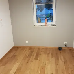 Pusty pokój z nową, jasną drewnianą podłogą, świeżo pomalowanymi szarymi ścianami i oknem z żaluzjami; puszka pianki montażowej na parapecie.