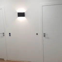 Dwie białe, nowoczesne drzwi wewnętrzne z poziomymi panelami, zamontowane w świeżo pomalowanym korytarzu, z czarnym, minimalistycznym kinkietem emitującym ciepłe światło i widocznymi puszkami...