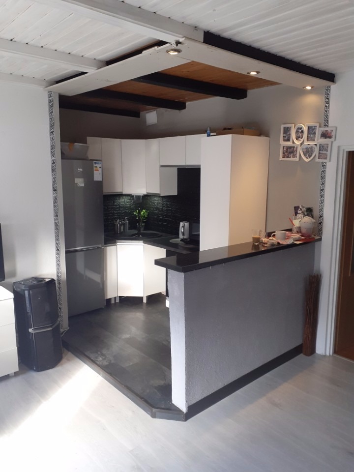 Jasna kuchnia z czarnym blatem, widoczne białe szafki, czarna ściana nad blatem, drewniany sufit i dekoracyjny pasek na ścianie.