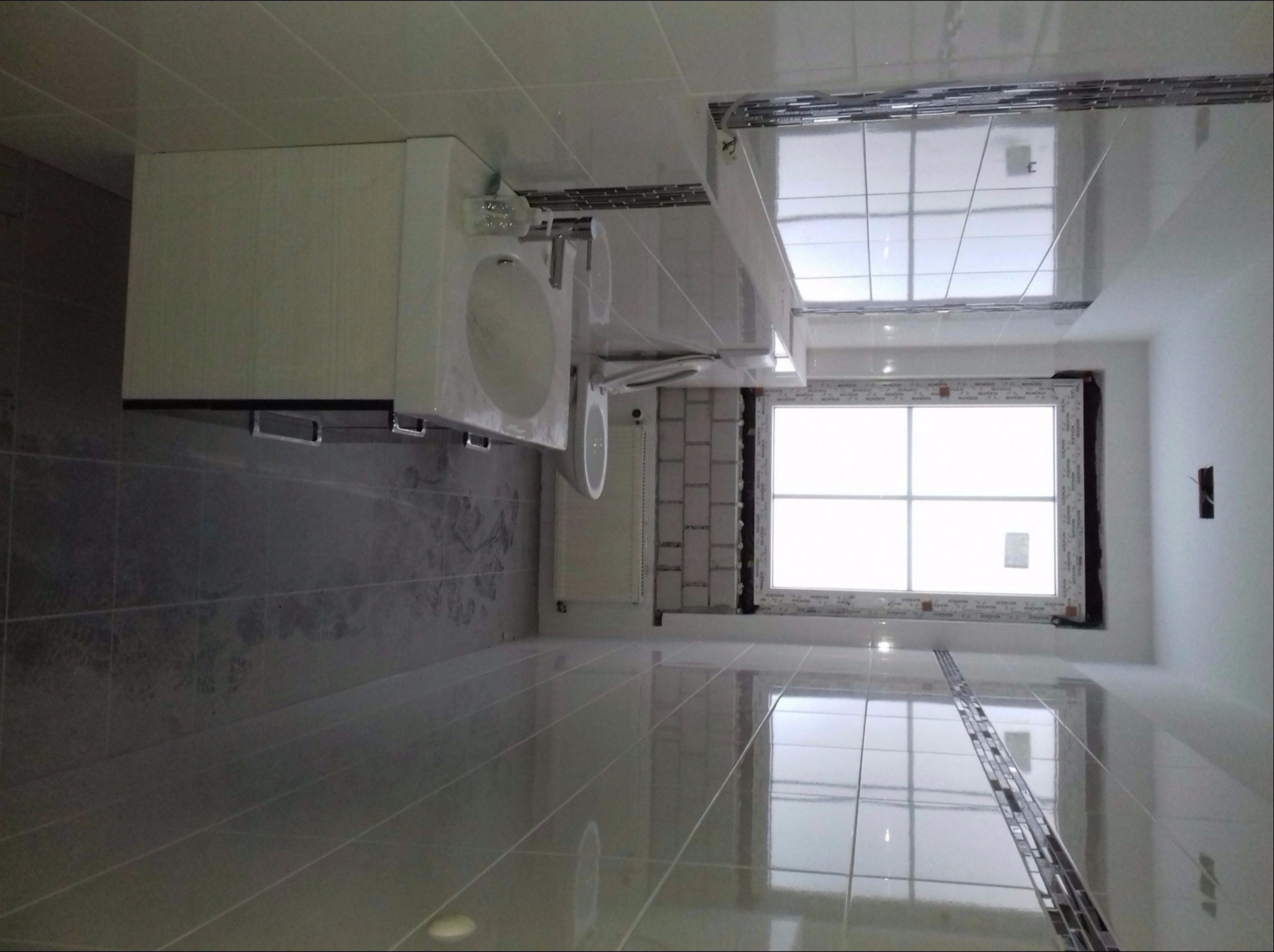 Nowoczesna łazienka z błyszczącymi, szarymi płytkami podłogowymi i ściennymi, białą szafką z umywalką i lustrem oraz oknem z białymi ramami, wykończona dekoracyjną listwą mozaikową.