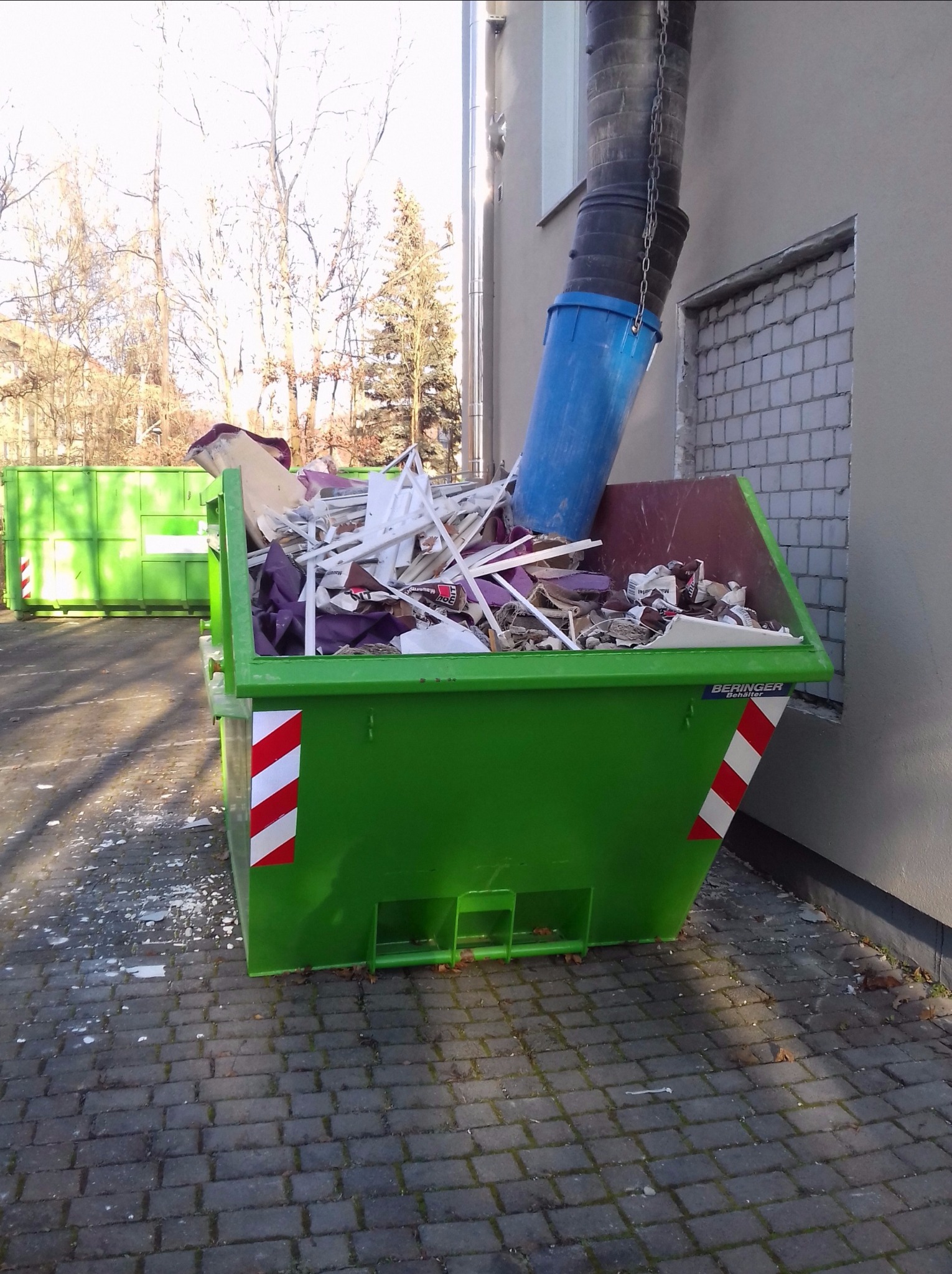 Zielony kontener na gruz wypełniony odpadami budowlanymi, z rurą zsypową w tle, na brukowanej powierzchni.