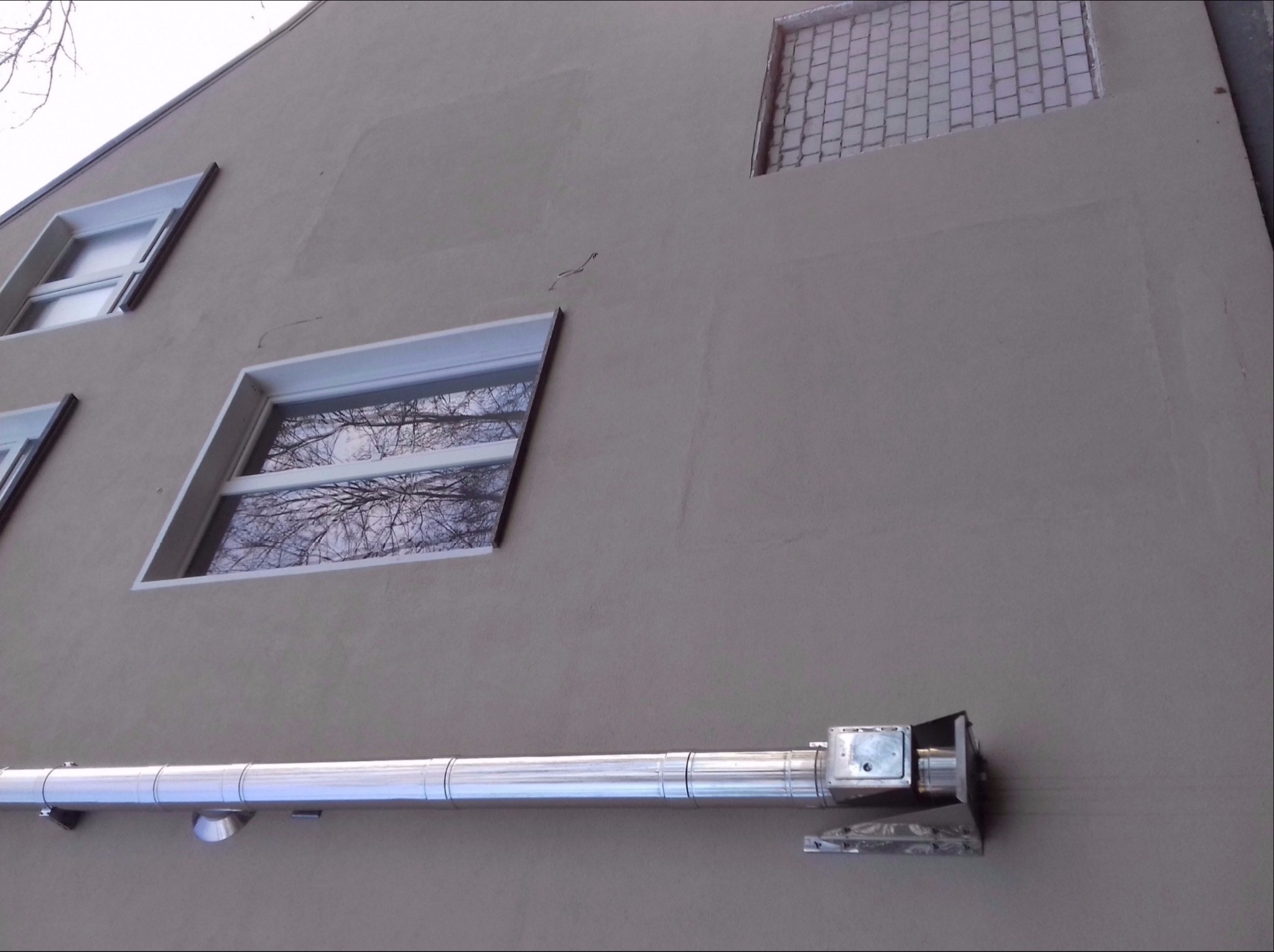 Szara elewacja budynku z widocznymi oknami i fragmentem systemu odprowadzania wody deszczowej, z zaznaczonymi obszarami po naprawach tynkarskich.