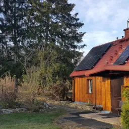 Dom jednorodzinny z panelami słonecznymi na czerwonym dachu, widok z ogrodu z drzewami iglastymi w tle.