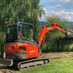 Pomarańczowa mini koparka Kubota KX027-4 w trakcie pracy, operator w kabinie, łyżka nabiera ziemię, zielona trawa i drzewa w tle, pagórkowaty krajobraz w oddali.