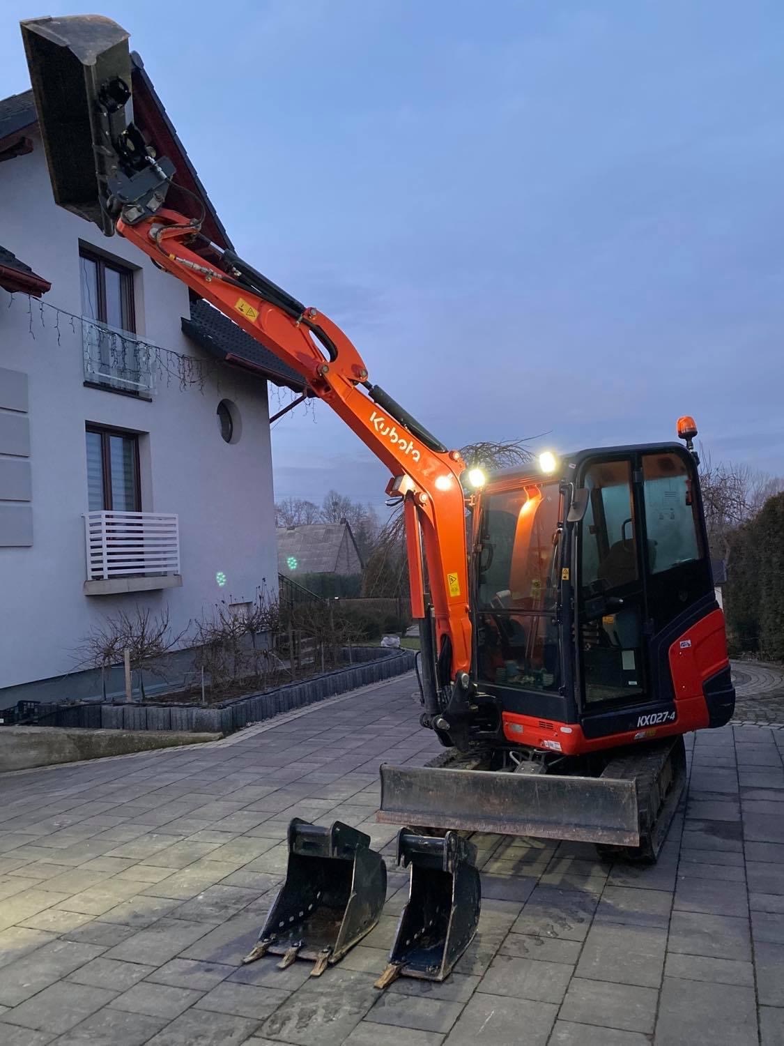 Pomarańczowa mini koparka Kubota KX027-4 z podniesionym ramieniem i łyżką, dodatkowe łyżki leżą na kostce brukowej przed maszyną, oświetlenie robocze włączone, dom w tle.