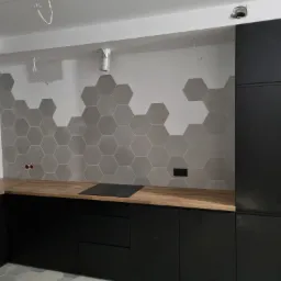 Wnętrze kuchni w trakcie wykańczania: czarne szafki z drewnianym blatem, ściana z szarymi heksagonalnymi płytkami, widoczne kable i puszki elektryczne, sufit z niedokończonym oświetleniem.