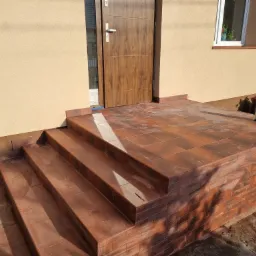 Kafelkowane schody wejściowe w kolorze terakoty prowadzące do brązowych drzwi wejściowych, z beżową elewacją budynku w tle.