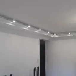 Białe ściany po malowaniu z dekoracyjną listwą sufitową z wbudowanymi punktowymi lampami LED oraz wiszący żyrandol z abażurem, widoczna osoba w tle.