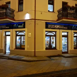 Fasada budynku Centrum Zaopatrzenia Ortopedyczno-Medycznego z balkonami i kwiatami, widoczny neon z logo firmy i wystawy sklepowe.