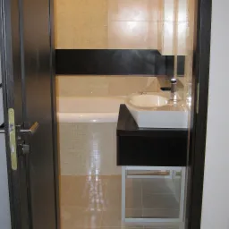 Widok przez otwarte drzwi na wykończoną łazienkę z beżowymi płytkami, wanną, umywalką nablatową na czarnej szafce z metalowym stelażem i chromowaną baterią.