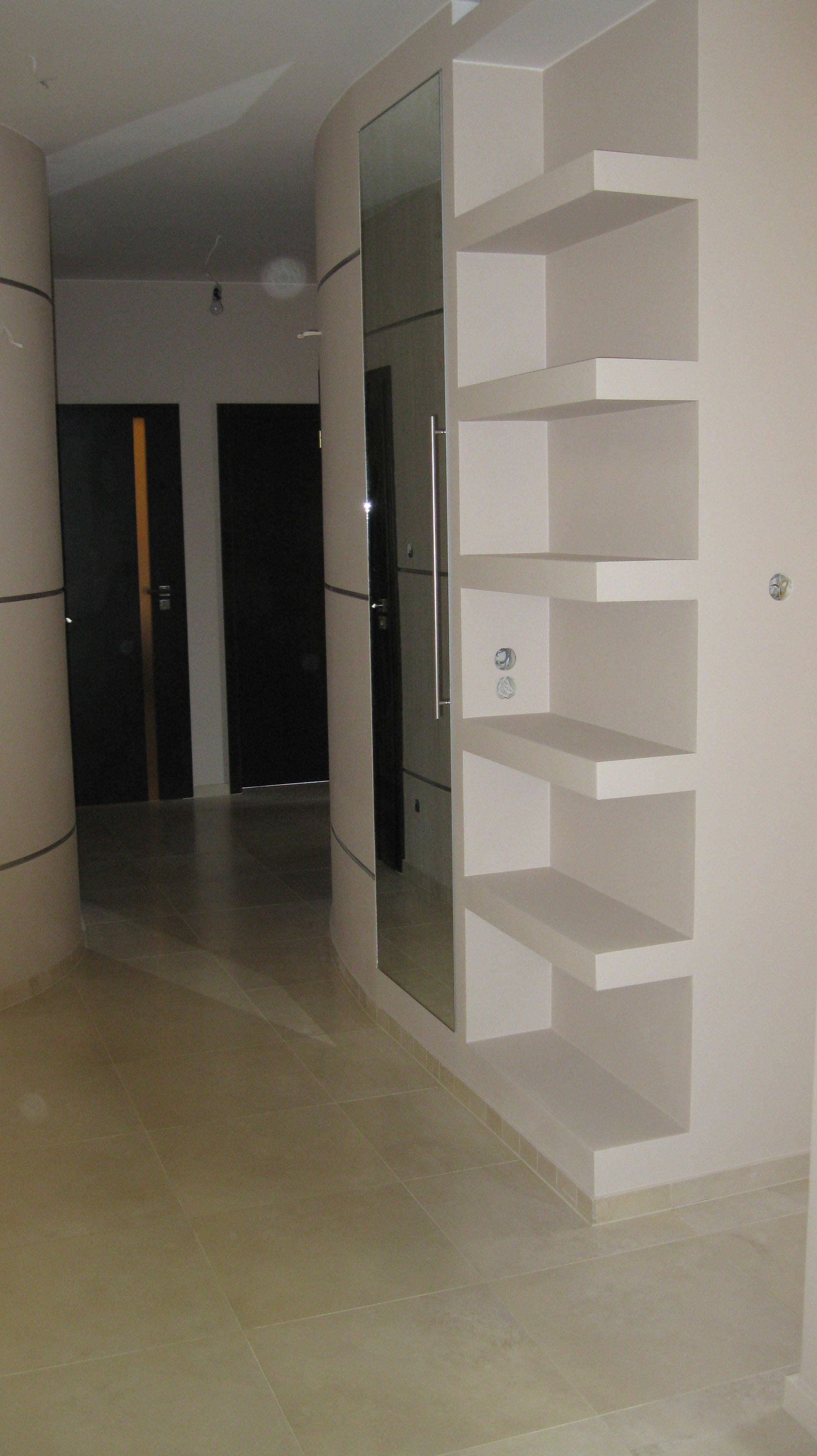 Wykończony korytarz z beżowymi płytkami podłogowymi, wbudowanymi półkami z oświetleniem punktowym i lustrem po lewej stronie, obok widoczne dwie ciemne drzwi.