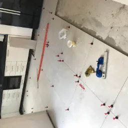 Układanie jasnych płytek podłogowych w kuchni, widoczne poziomice, przyssawka do płytek, krzyżyki dystansowe i system poziomowania płytek z czerwonymi klinami.