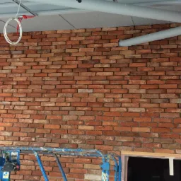 Wnętrze w trakcie prac wykończeniowych: ściana z czerwonej cegły, częściowo zakryta rusztowaniem, obok ściana z płyt gipsowo-kartonowych przed szpachlowaniem, widoczne rury pod sufitem.