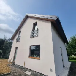 Nowoczesny dom szkieletowy z jasną elewacją, małymi balkonami z metalowymi barierkami i widocznymi elementami wykończeniowymi wok&oacute;ł budynku.