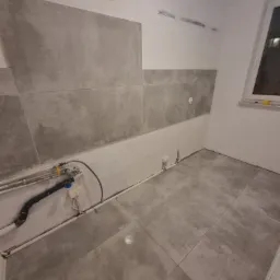 Szara, częściowo wykończona łazienka z ułożonymi płytkami podłogowymi i częściowo położonymi płytkami ściennymi, widoczne wystające rury i instalacje elektryczne.