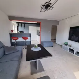 Nowoczesny salon z szarą sofą, czarnym stolikiem kawowym na metalowej nodze, barkiem z czerwonymi hokerami oraz telewizorem na ścianie, widok na kuchnię w tle.