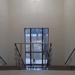 Klatka schodowa z metalową balustradą i okienkiem z widokiem na las, ściany wykończone w jasnym odcieniu, widoczna lampa sufitowa.