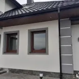 Jasna elewacja domu z szarymi detalami architektonicznymi wokół okien i na narożnikach. Widoczne dwa okna z brązowymi ramami i fragment dachu z ciemną dachówką. Na pierwszym planie schody wejściowe...