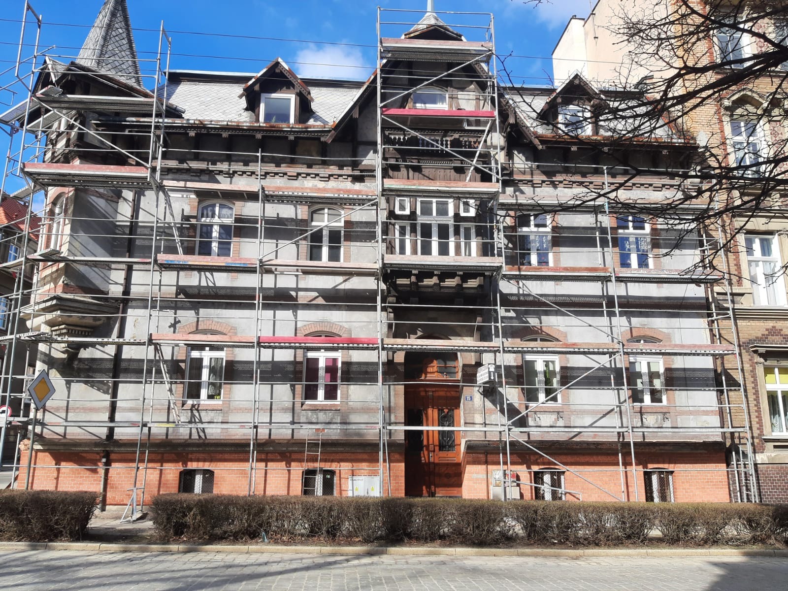 Renowacja kamienicy z rusztowaniem, widoczna część elewacji z oknami i detalami architektonicznymi, krzewy przed budynkiem, błękitne niebo w tle.