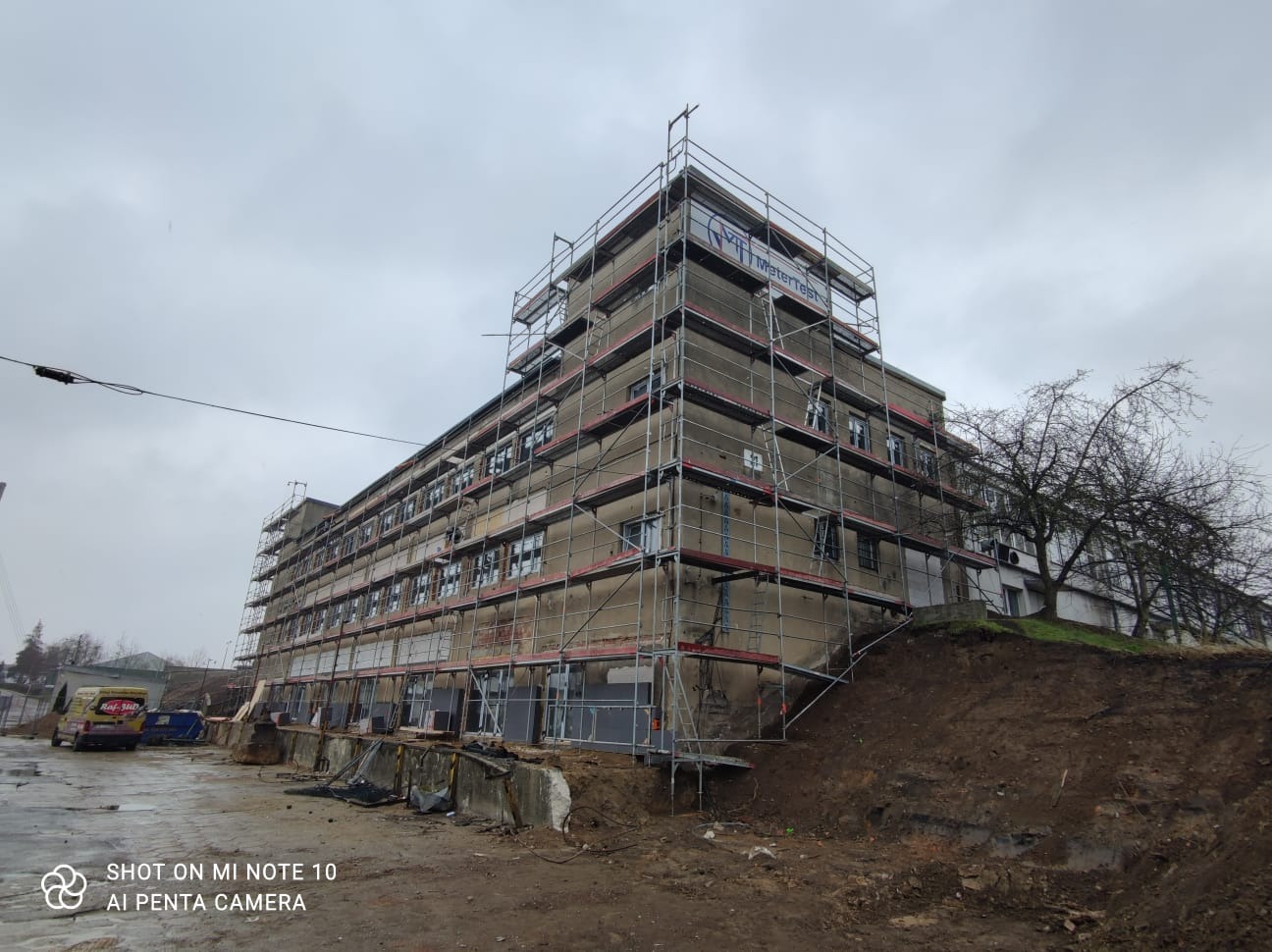 Budynek w trakcie remontu, otoczony rusztowaniami, z widocznym banerem firmy na szczycie, na tle pochmurnego nieba.