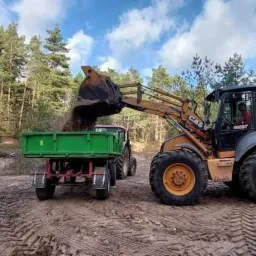 Żółto-czarna koparko-ładowarka Case 580ST nabiera ziemię i wysypuje do zielonej przyczepy ciągniętej przez czerwony traktor na tle lasu i błękitnego nieba.